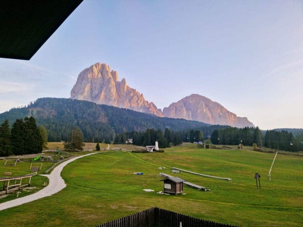 Where To Stay In The Dolomites: 9 Best Areas (+ The Worst!) 2 A scenic view of grassy fields leading to a majestic mountain range bathed in warm sunlight. The foreground shows meandering paths and small structures, with dense forests at the base of towering rocky peaks under a clear sky.