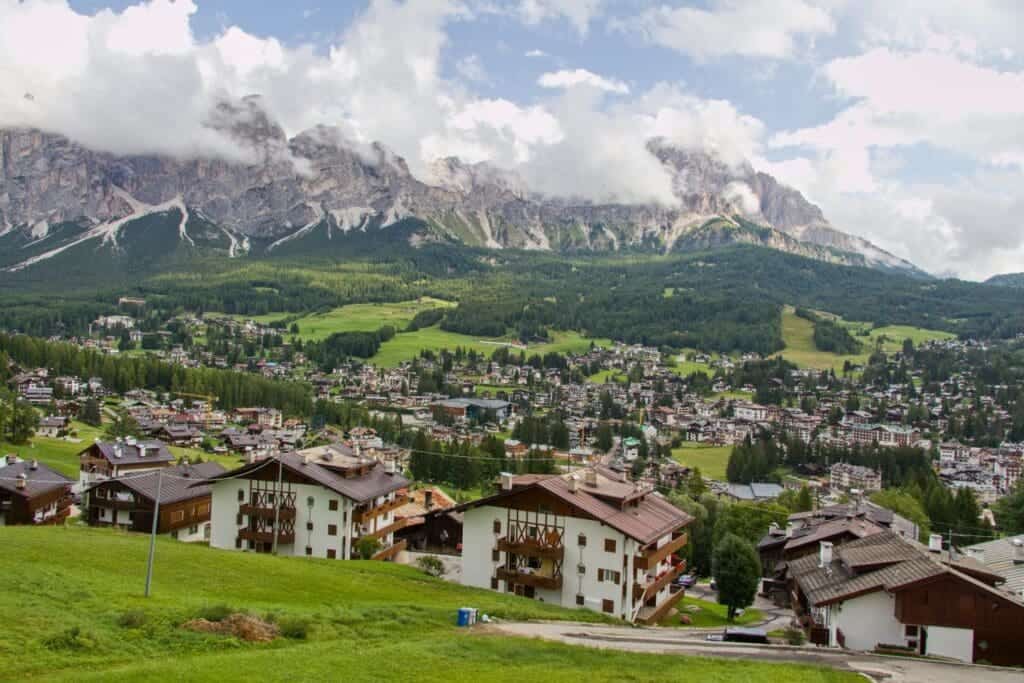 A scenic view of a mountainous landscape with a cloudy sky, showcasing a town nestled in a valley. Traditional Alpine houses with wooden balconies are surrounded by lush green fields and dense forests, with majestic mountains looming in the background.