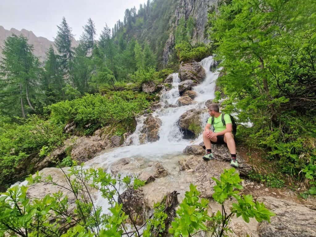 Cool Things To Do In The Dolomites: From Hiking to Flying 12 A person in a green shirt and shorts sits by a small waterfall in a lush, mountainous landscape. Surrounding them are green bushes and tall evergreen trees. The sky is overcast, and the scene is calm and serene.
