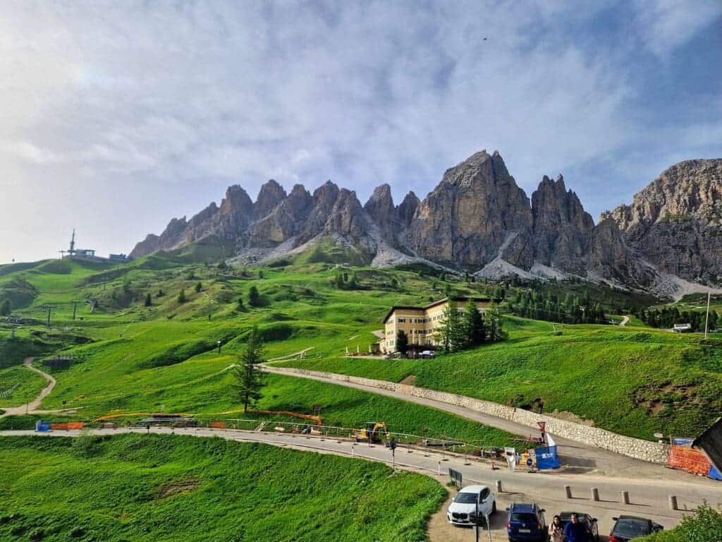 Green hills with a winding road lead to a yellow building, set against dramatic, jagged mountain peaks under a blue sky with scattered clouds. Several parked cars and construction equipment are visible in the foreground.