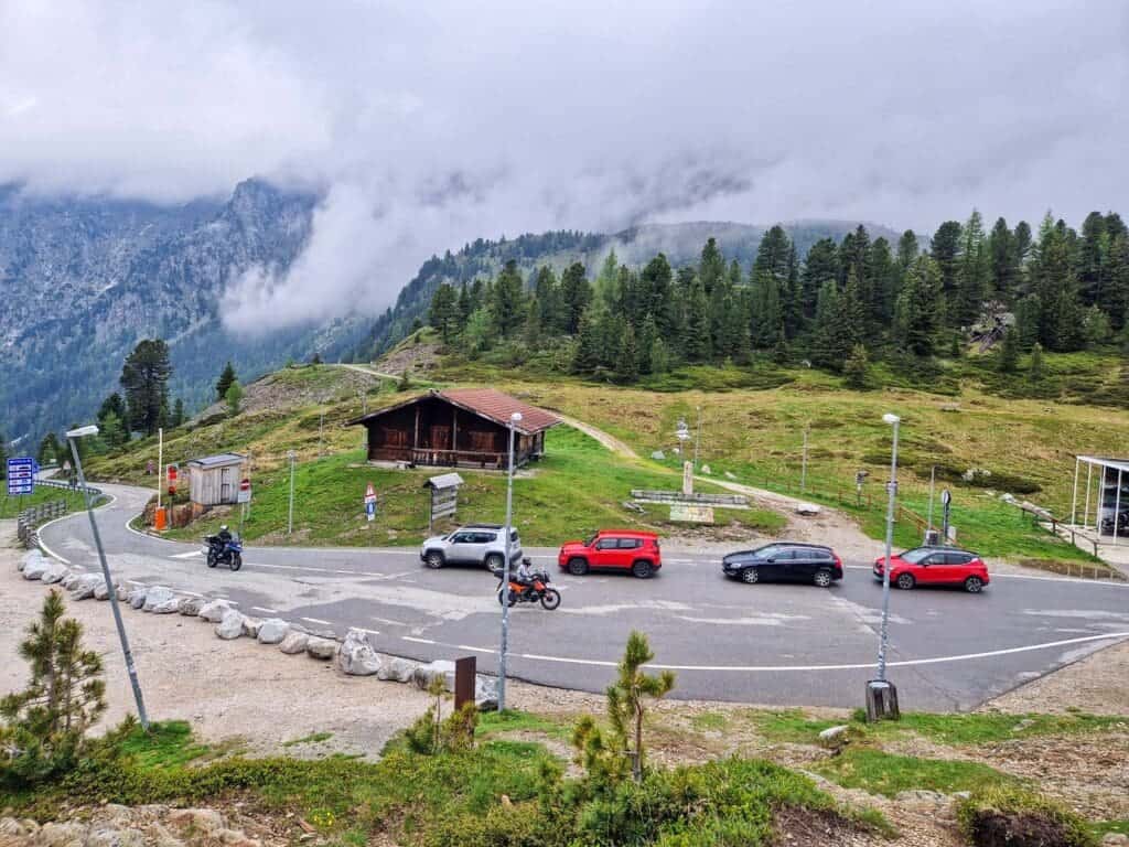 A winding mountain road with several cars and motorcycles parked beside a wooden cabin, surrounded by green hills, pine trees, and misty mountains in the background.