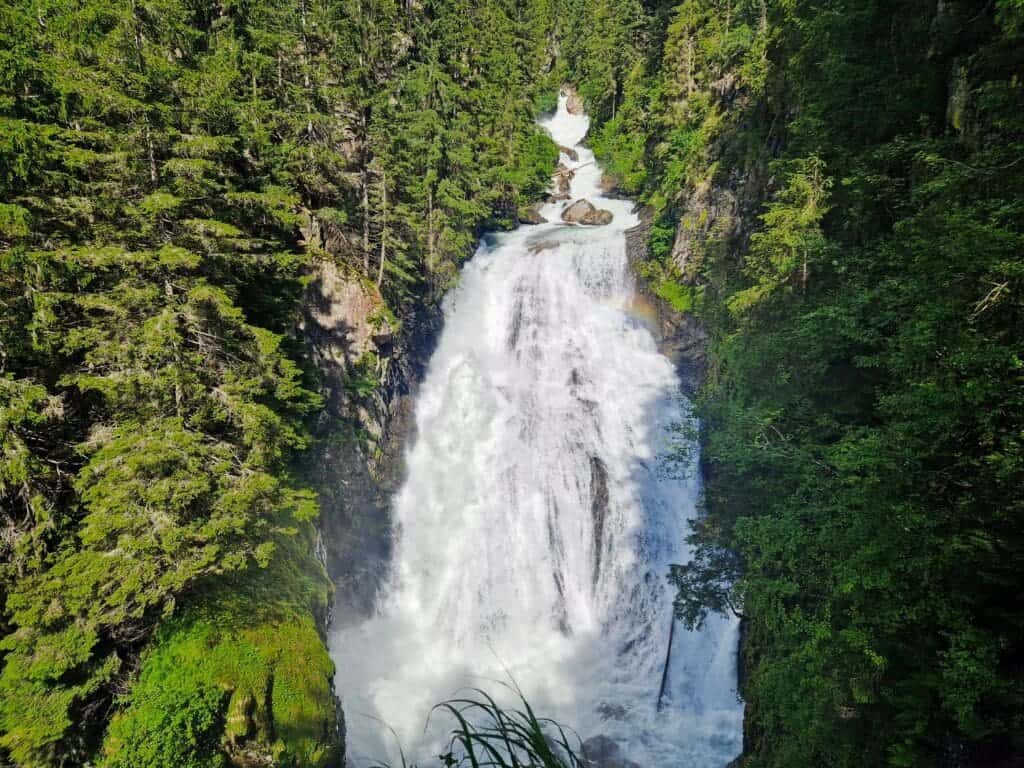 Cool Things To Do In The Dolomites: From Hiking to Flying 13 A scenic view of a waterfall cascading down rocks surrounded by lush, green forest. Sunlight highlights the water, creating a faint rainbow in the mist. Dense trees frame the waterfall, adding to the serene, natural beauty of the scene.