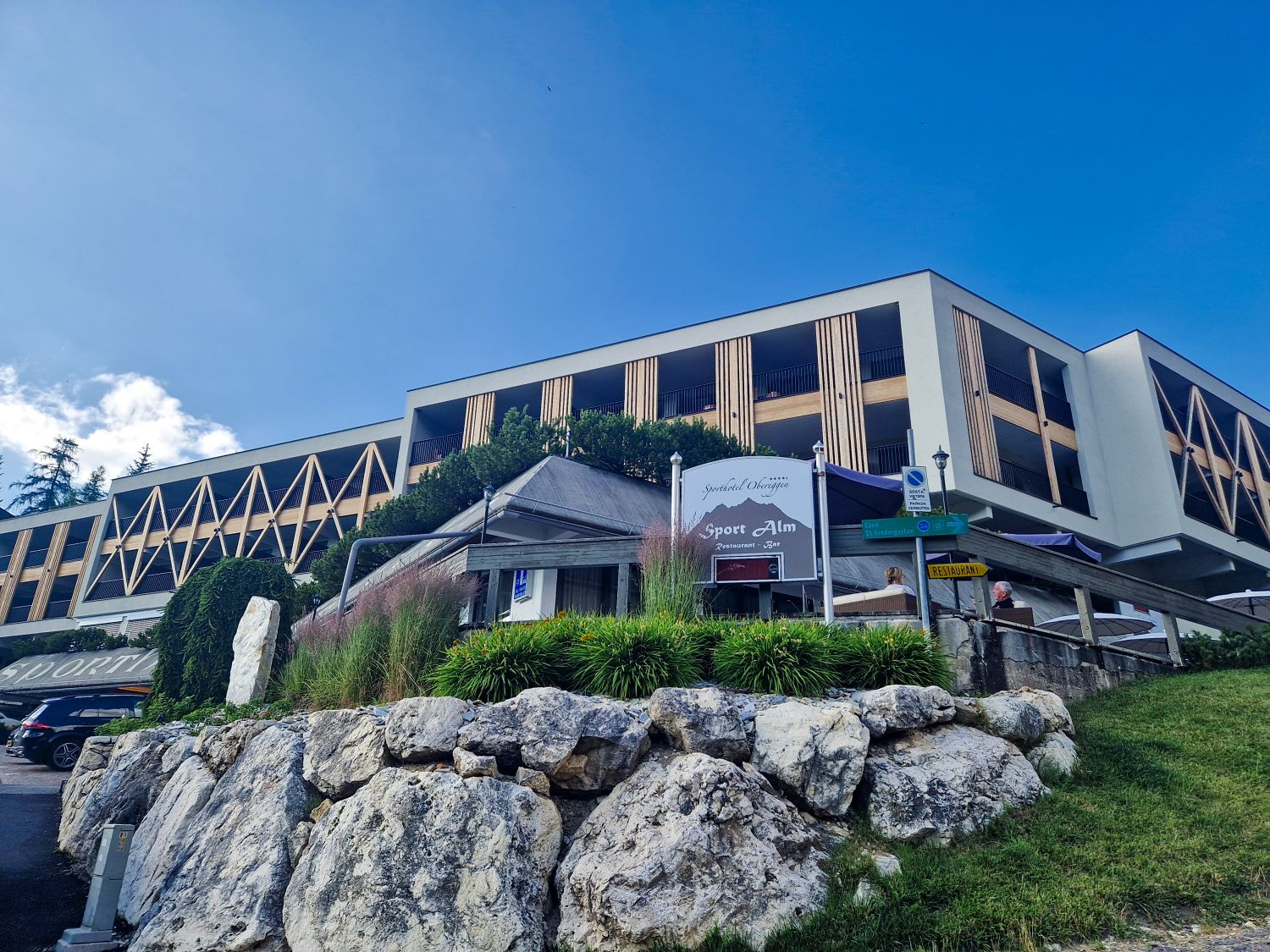 A modern hotel building with geometric wooden accents sits on a hill, surrounded by greenery and large rocks. Signs in front indicate directions and the name "Sport Alm." The sky is bright and clear.