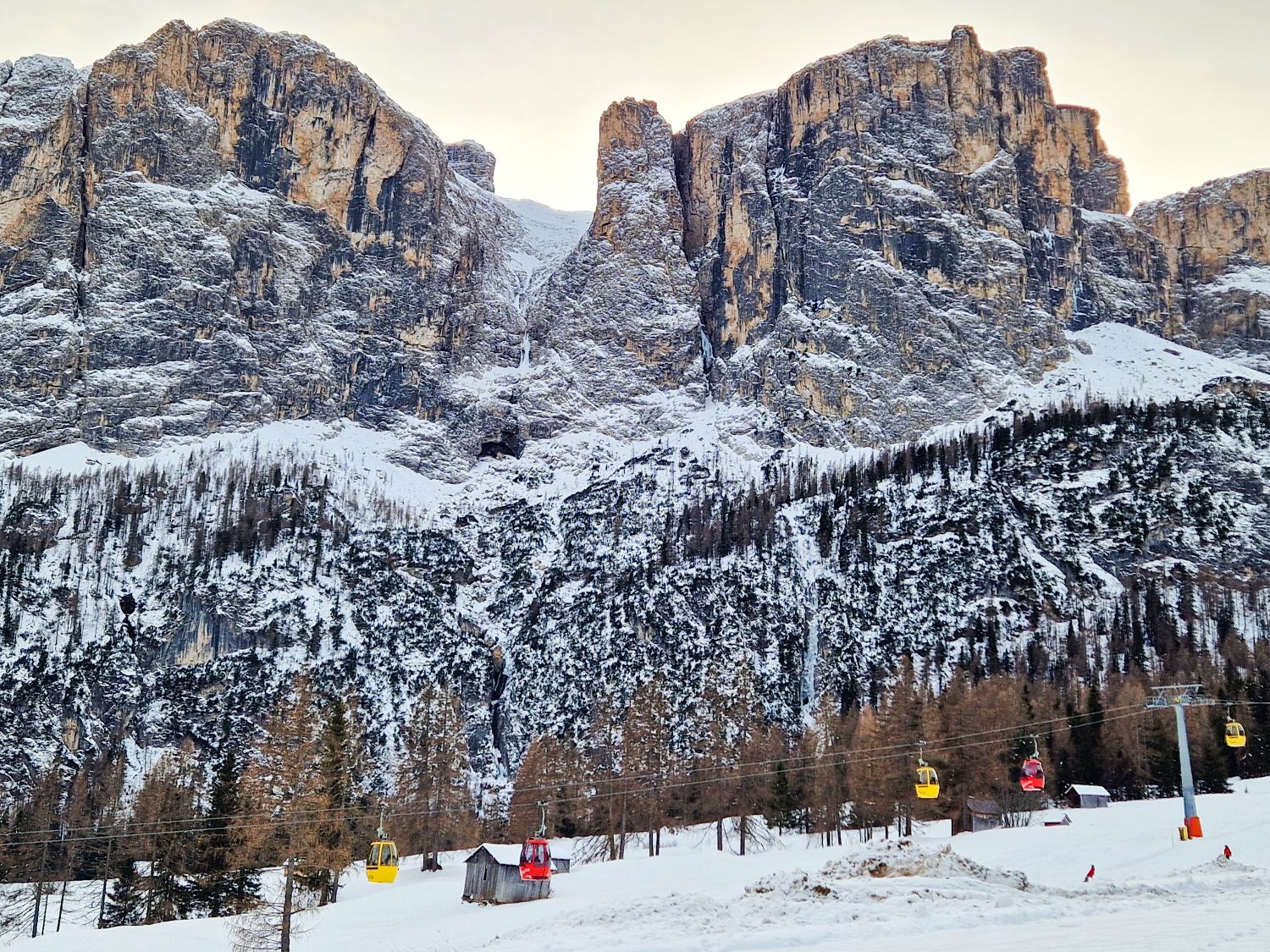 Things To Do In The Dolomites In Winter: Ski And No Ski 13 A dramatic snow covered mountain cliff rises above a winter ski area with dark rock faces dusted in fresh snow. In the foreground colorful gondola cars hang from a cable line over a white slope with small wooden huts and sparse trees below.