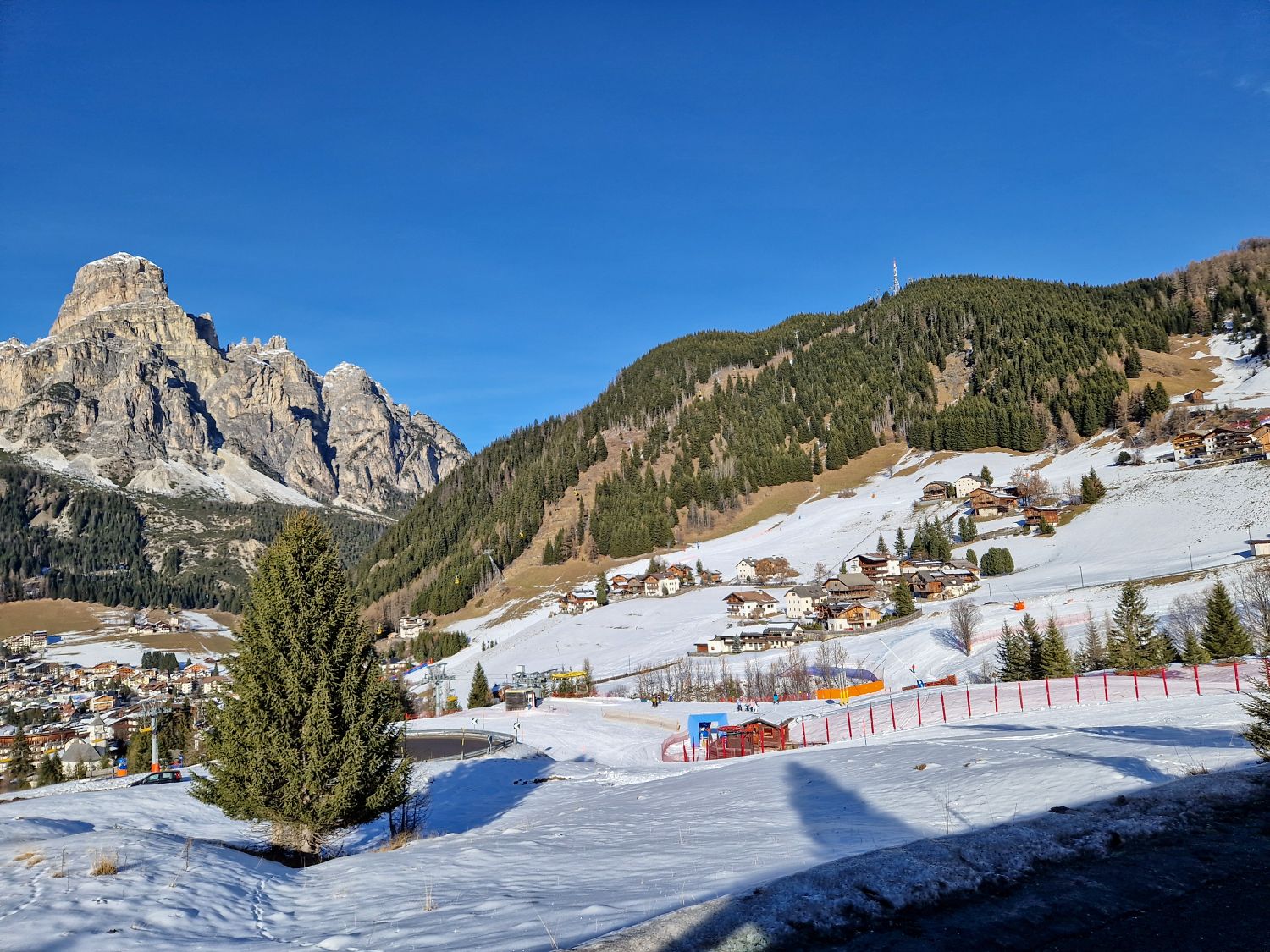 Where to stay in the Dolomites for skiing 7 A snowy alpine village sits on rolling hills beneath a clear blue sky with evergreen forests and rugged mountain peaks in the background. Ski slopes with red safety fencing wind through the scene alongside scattered chalets trees and lift infrastructure creating a bright winter landscape.