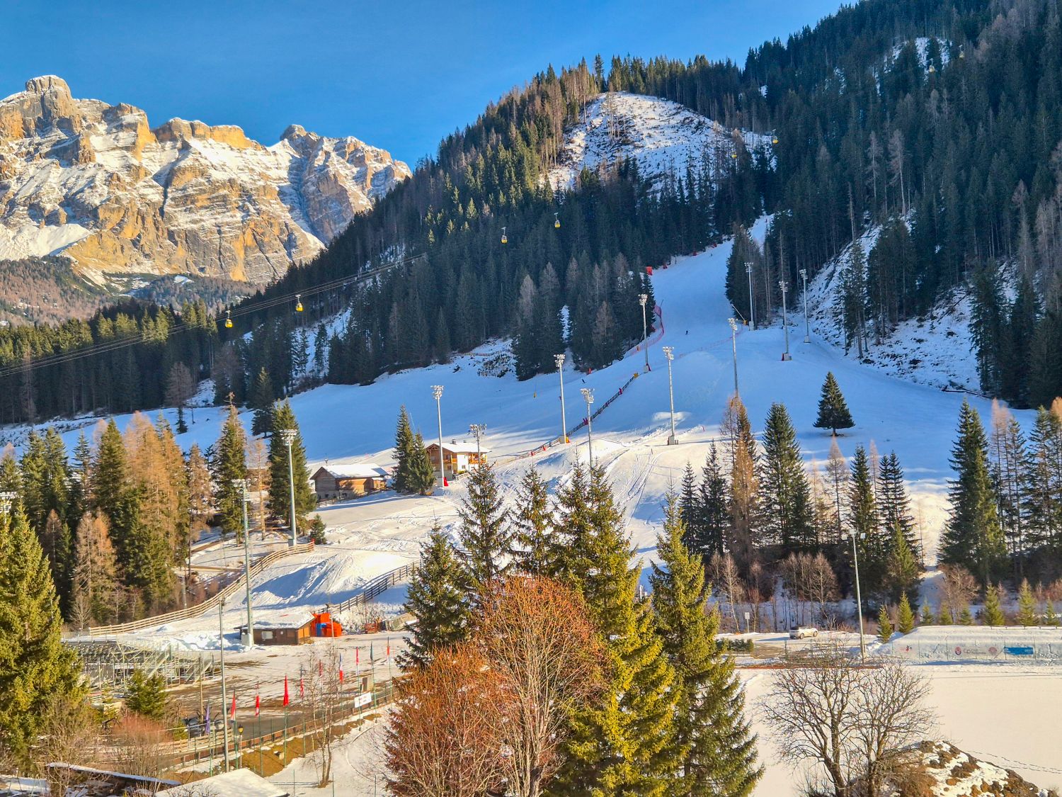 Skiing in the Dolomites for the first time 9 A sunlit ski area sits in a forested mountain valley with groomed slopes cutting through evergreen trees. Yellow gondola cars glide across the hillside while ski lifts buildings and light poles line the snow covered runs beneath rugged rocky peaks.