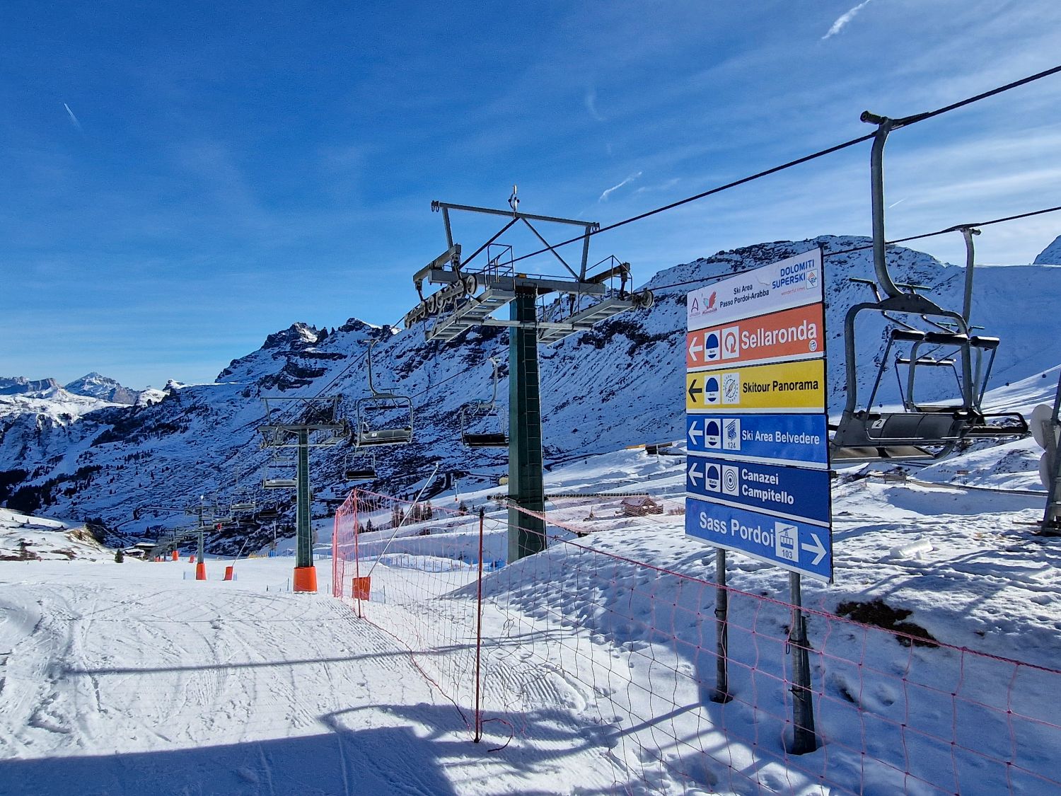 Skiing in the Dolomites for the first time 10 Snowy alpine ski area with a chairlift running overhead and rugged mountain peaks in the background under a clear blue sky. In the foreground a large directional sign reads Dolomiti Superski and shows arrows for Sellaronda, Skitour Panorama, Ski Area Belvedere, Canazei Campitello, and Sass Pordoi near Passo Pordoi Arabba. The image helps orient skiers by showing trail connections and lift access points at a mountain junction.