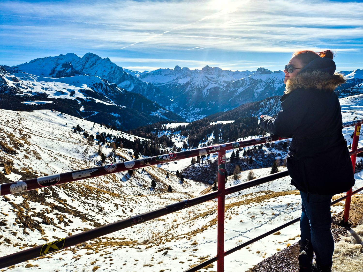 Dolomites in Winter: Weather, Skiing, Hiking, and more 18 Person in a winter jacket stands at a mountain viewpoint looking out over a wide snow covered valley and jagged alpine peaks under a bright sky. A red metal railing lines the overlook while forested slopes and winding terrain drop away below. The image captures a quiet moment of scale and perspective in a high mountain landscape.