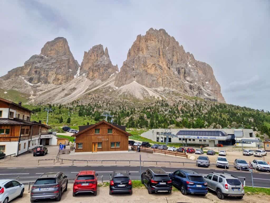 A scenic view of towering rocky mountains under a cloudy sky. In the foreground, a row of parked cars lines a road in front of wooden buildings. Green trees dot the landscape, adding contrast to the rugged mountain backdrop.