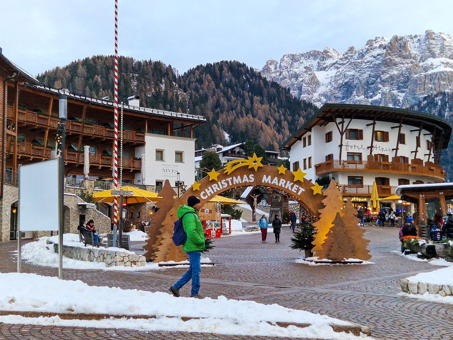 Dolomites in Winter: Weather, Skiing, Hiking, and more 5 A snowy Christmas market in Selva di Val Gardena at the beginning of December, surrounded by traditional Alpine buildings and the snow covered Dolomites. A large wooden arch reading "CHRISTMAS MARKET" welcomes visitors walking through the cobblestone village square.