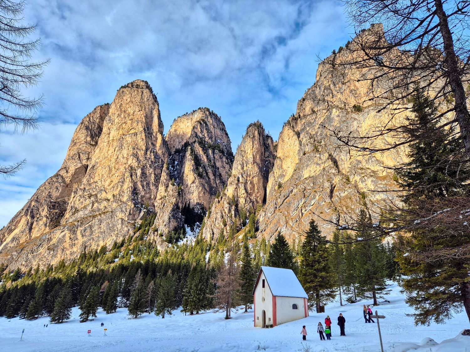 Dolomites in Winter: Weather, Skiing, Hiking, and more 17 Vallunga near Selva in the Dolomites in December. Dramatic golden rock cliffs rise above a snow covered forest and a small chapel under a bright blue winter sky.