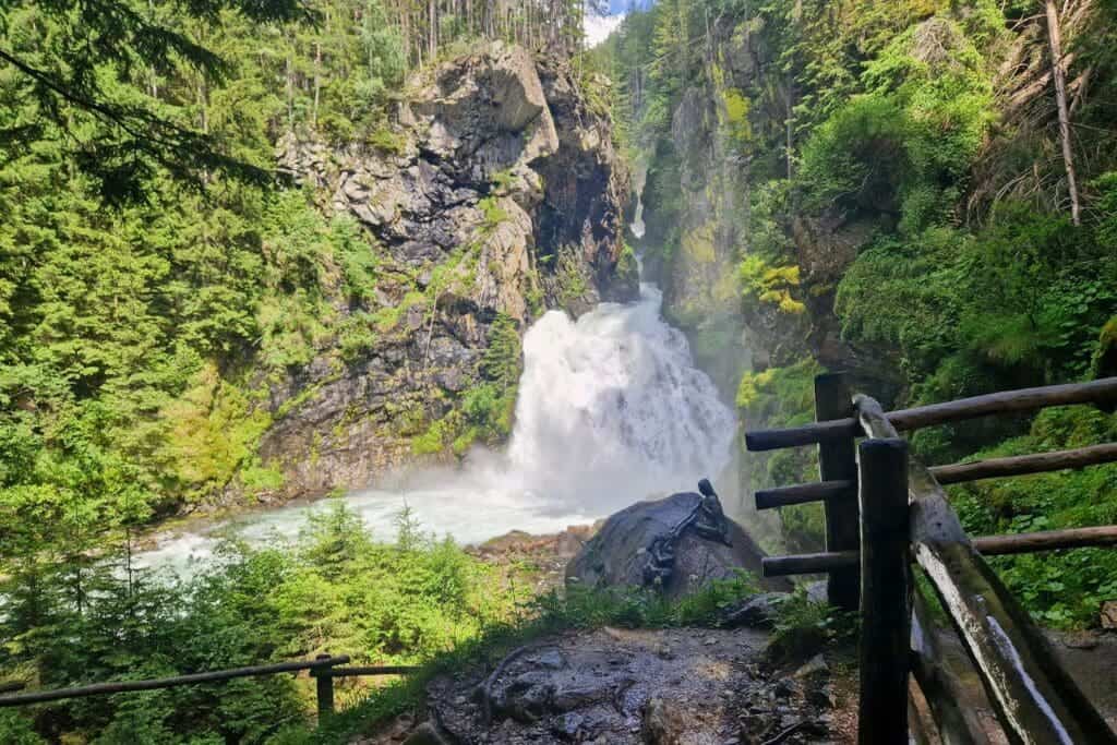 Cool Things To Do In The Dolomites: From Hiking to Flying 14 A scenic view of a waterfall cascading down a rocky cliff, surrounded by lush greenery and trees. A wooden fence lines a pathway in the foreground, leading to the waterfall. Sunlight filters through the trees, illuminating the scene.