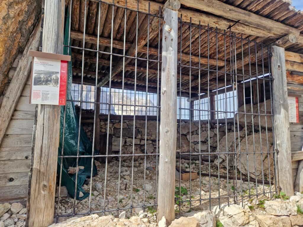 A rustic wooden and stone shelter with a metal gate stands on a rocky terrain. Informational signs are placed on the wooden beams. The shelter's roof is made of overlapping wooden planks, and the interior appears empty.