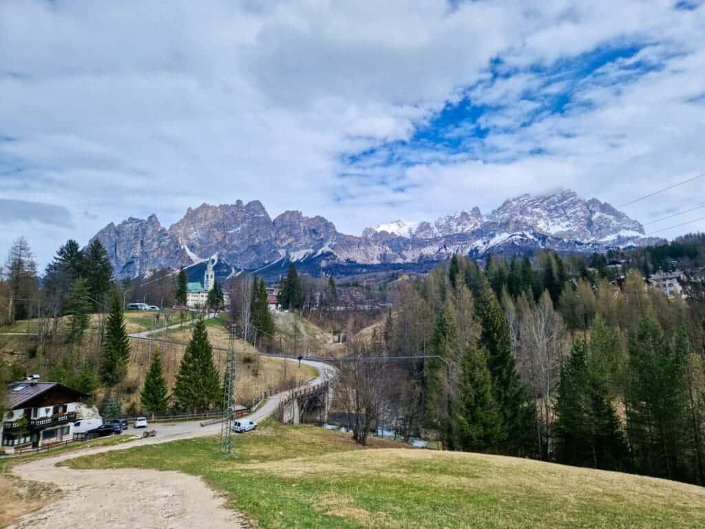 A scenic landscape features a winding road leading through a hilly area with sparse trees. In the background, rugged snow-capped mountains rise under a partly cloudy sky. A few houses and a small church are nestled among the trees.