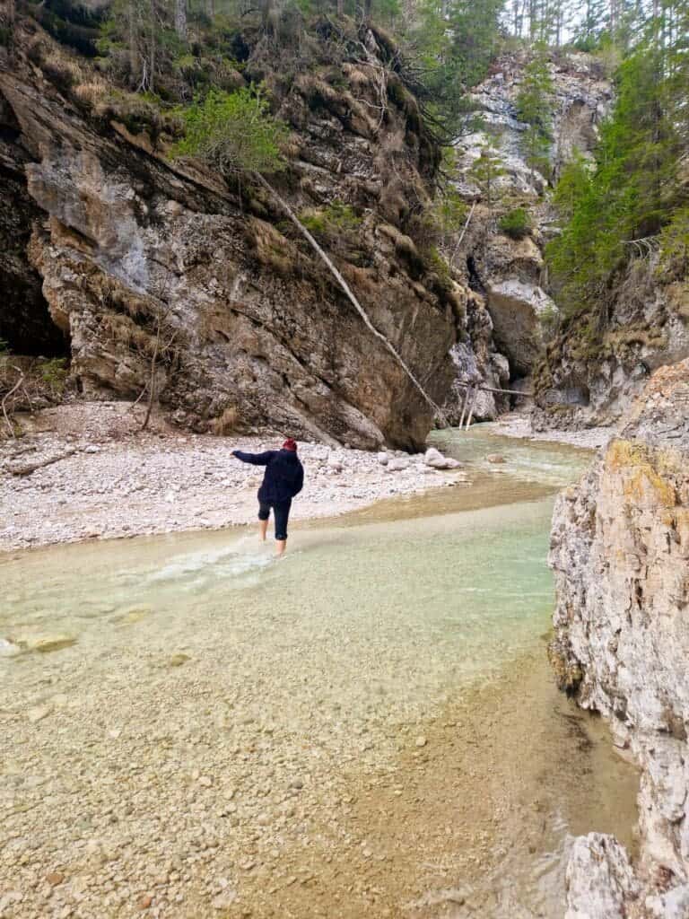 Dolomites In April: Perfect 2-Day Itinerary + What To Expect 43 A person wearing a dark jacket wades through a shallow, clear stream in a rocky canyon. Steep cliffs and green trees surround the area, creating a serene natural landscape. The person appears to be carefully balancing on the rocky bed.