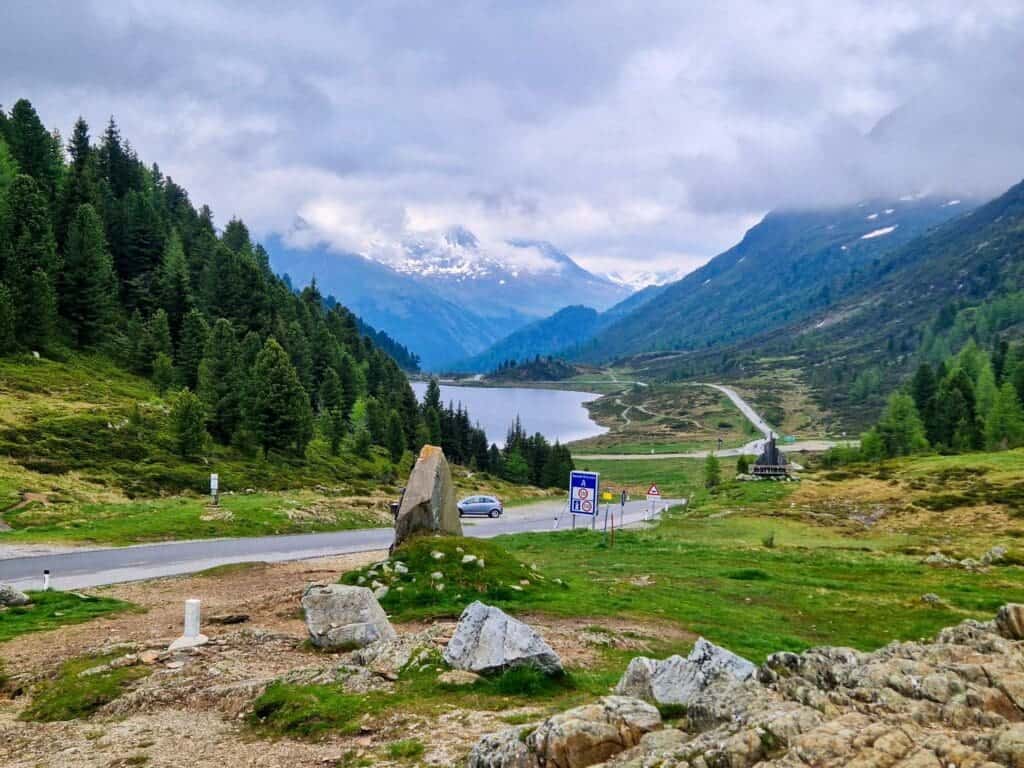 Mountain landscape with a winding road cutting through lush green hills and leading towards a distant lake surrounded by snow-capped peaks. A few scattered rocks and a road sign are in the foreground, under a cloudy sky.