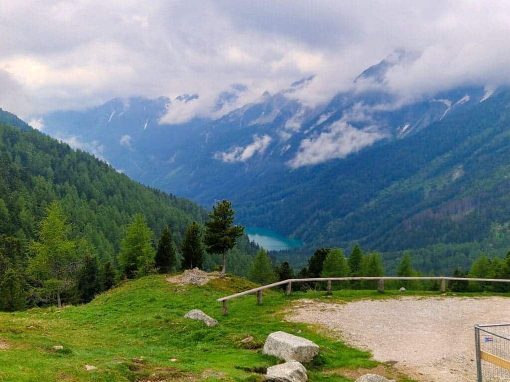 Mountain landscape with lush green forests and a valley containing a small, turquoise lake. The sky is cloudy, with mist partially covering the rugged peaks. A wooden fence and pathway are in the foreground.