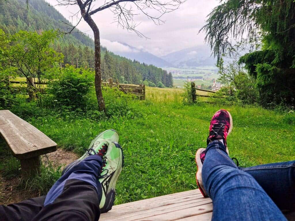 Where To Stay In The Dolomites: 9 Best Areas (+ The Worst!) 4 Two people relax on a wooden bench, overlooking a scenic valley with green fields and distant mountains. They wear athletic shoes—one green and black, the other pink and black—and jeans, surrounded by lush trees and a cloudy sky.