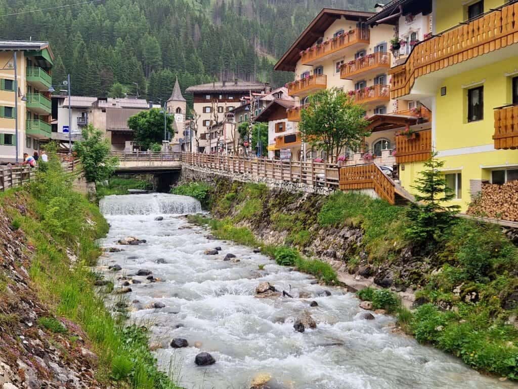 A scenic mountain village with a flowing river lined by colorful buildings and wooden balconies. A small bridge crosses the river, with lush green trees and mountains in the background on a bright day.