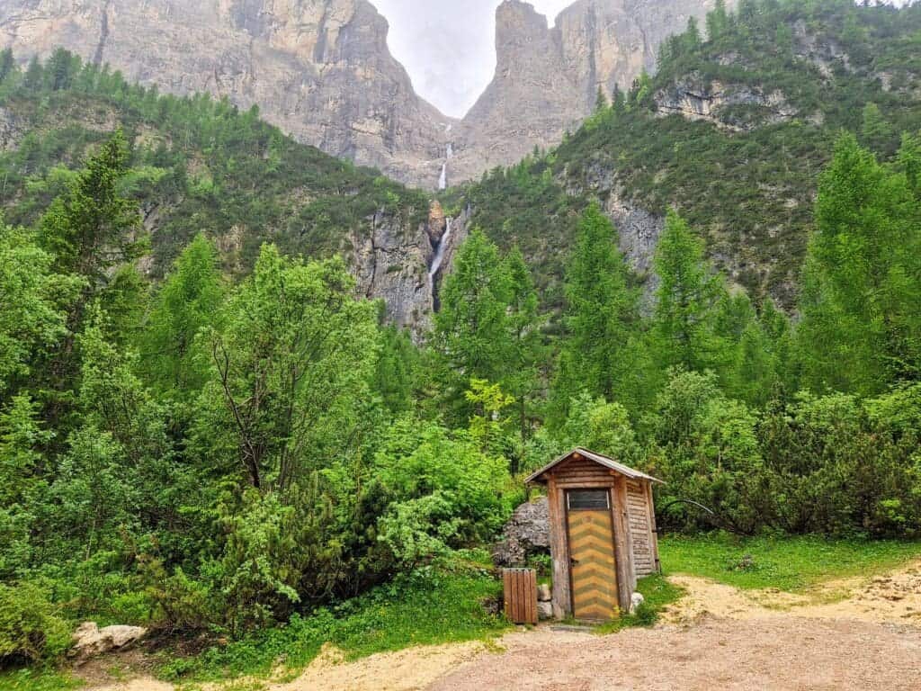 A small wooden hut with a zigzag-patterned door sits amidst lush green trees and shrubs. In the background, a tall waterfall cascades down a rocky mountain face under a cloudy sky. A path leads to the hut.