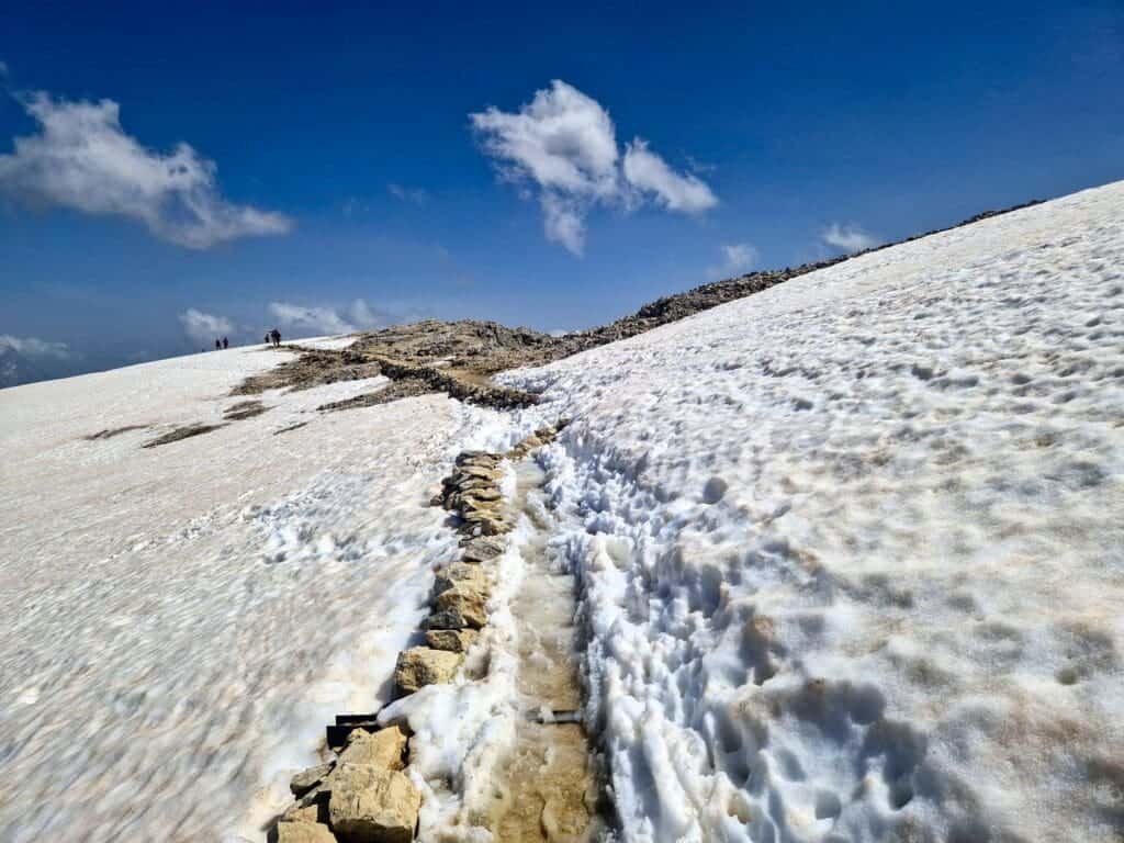 A narrow, rocky path winds through a snow-covered mountain slope under a clear blue sky. Few clouds are scattered above, and small figures can be seen in the distance along the path. Sparse rocks are visible along the snow.
