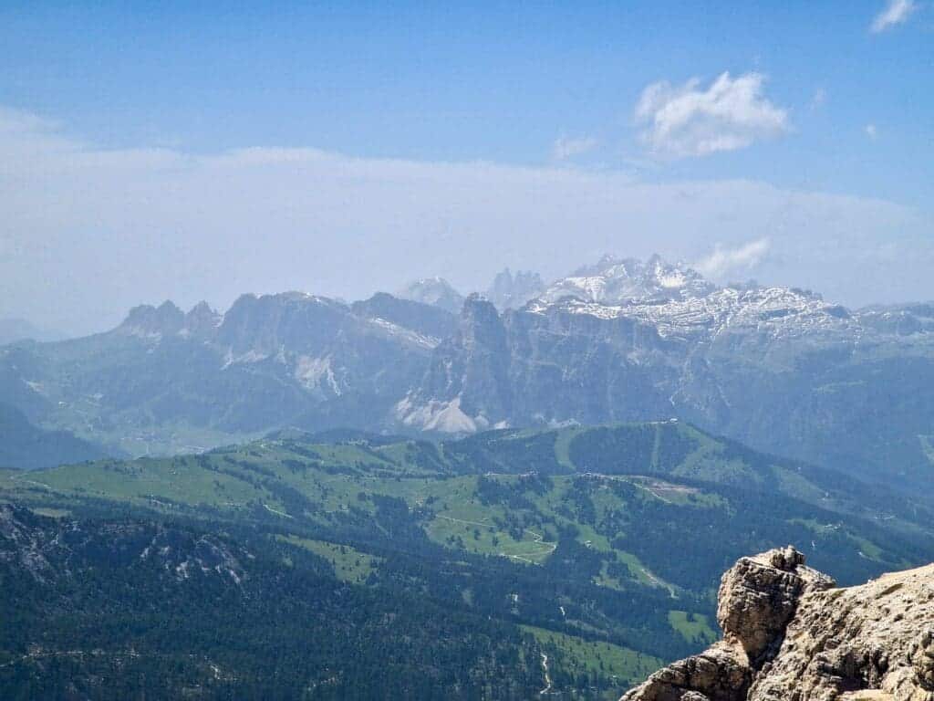 A panoramic view of rugged mountain ranges with snow-capped peaks under a clear blue sky. The foreground features rocky outcrops, and the middle ground shows rolling green hills and forests.
