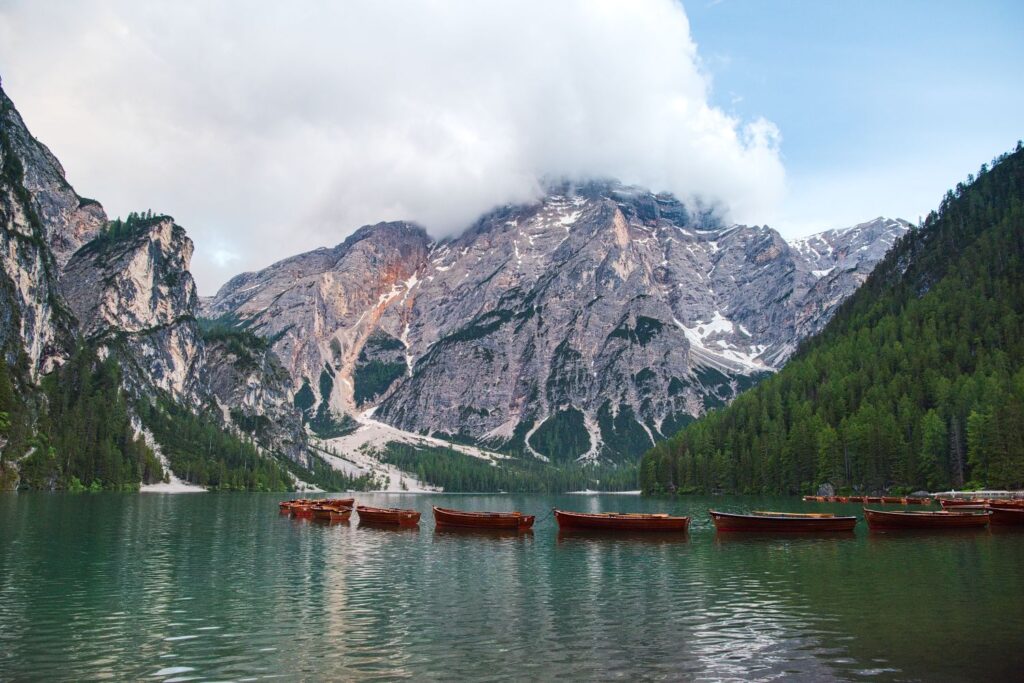 A serene mountain lake with several wooden boats floating on the calm water. The surrounding landscape features tall, rocky mountains partially covered with clouds and dense green forests along the shoreline.
