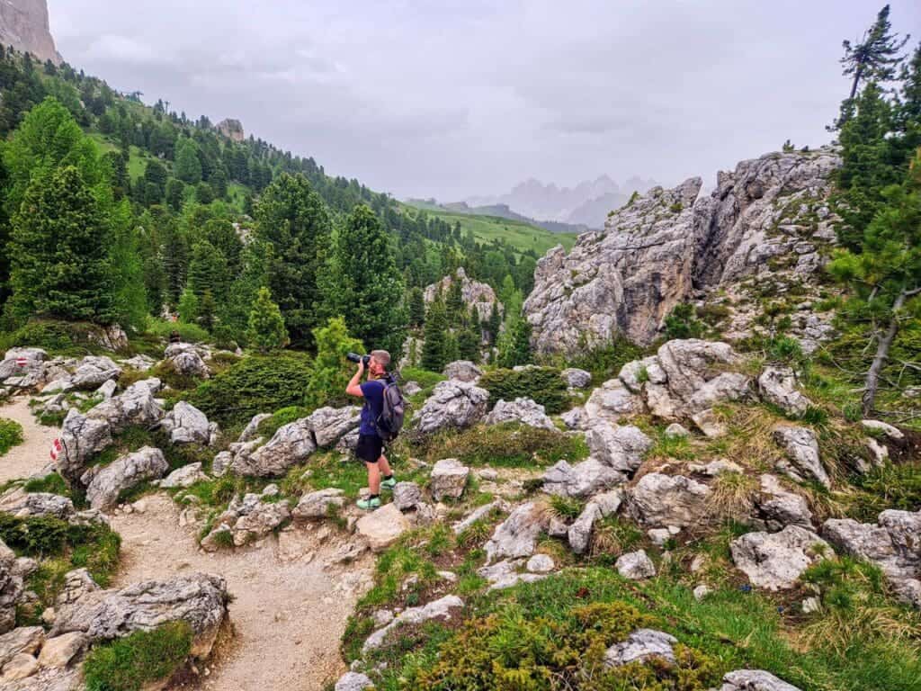 A person stands on a rocky path, taking photos with a camera in a mountainous landscape. The scene features lush green trees, shrubs, and distant rolling hills under a cloudy sky.