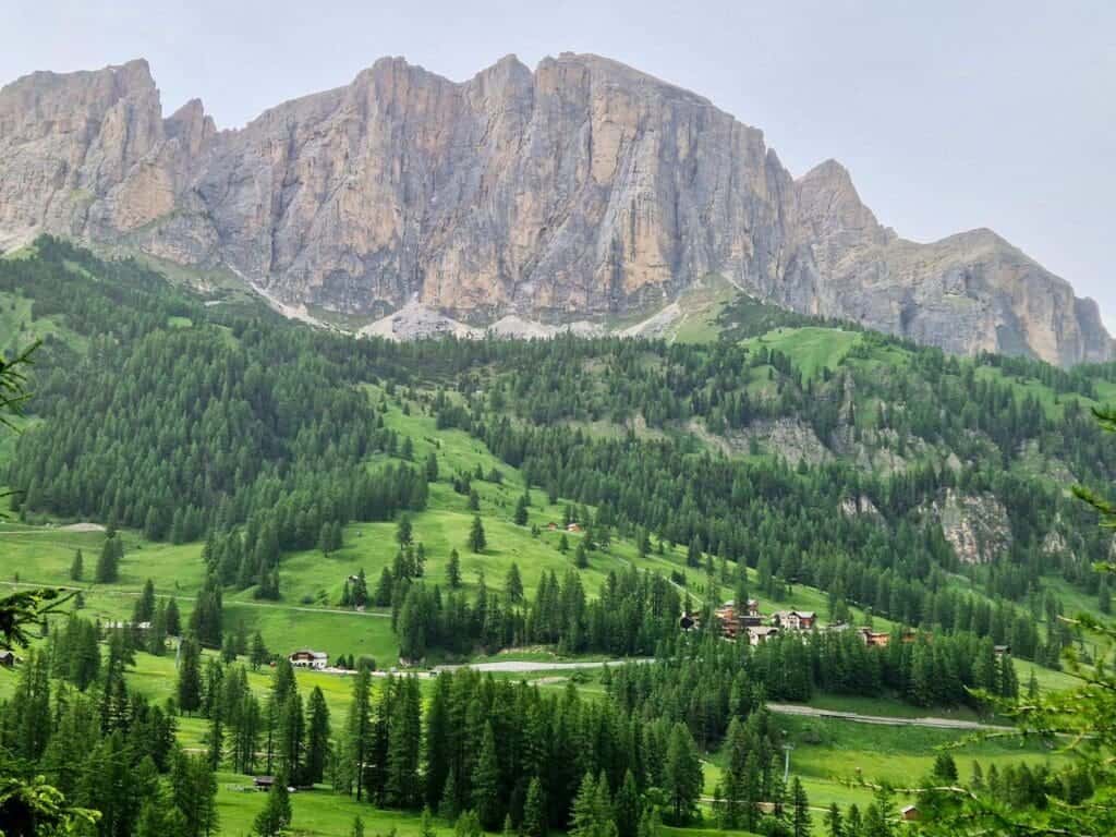 A scenic view of a lush green landscape with a rugged mountain range in the background. Pine trees cover the rolling hills, and a small cluster of buildings is visible in the valley below. The sky is overcast, casting a soft light over the scene.