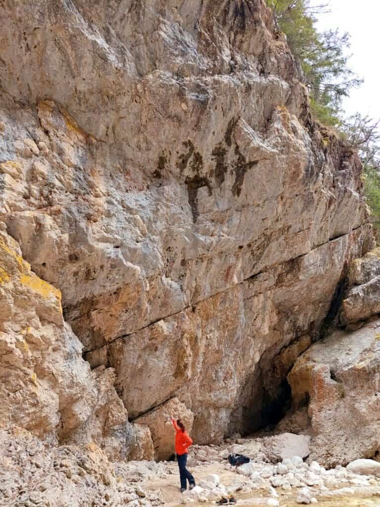 Dolomites In April: Perfect 2-Day Itinerary + What To Expect 45 A person in a red shirt and blue pants stands beneath a large, steep rock face, gesturing upward. The rocky terrain is rugged, with scattered stones at the base. Sparse greenery is visible at the top right.