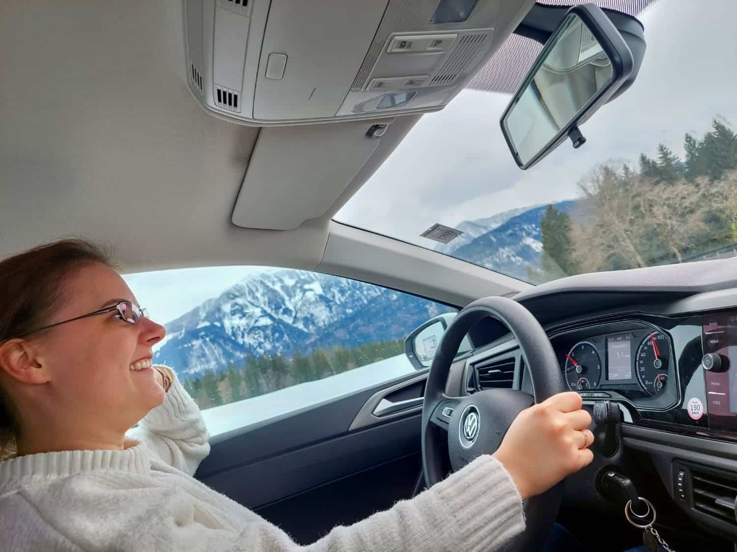 Dolomites In April: Perfect 2-Day Itinerary + What To Expect 56 A person wearing glasses and a white sweater is driving a car with a smile. The interior of the car is visible, including the dashboard and rearview mirror. Snow-covered mountains and trees are seen outside the window.