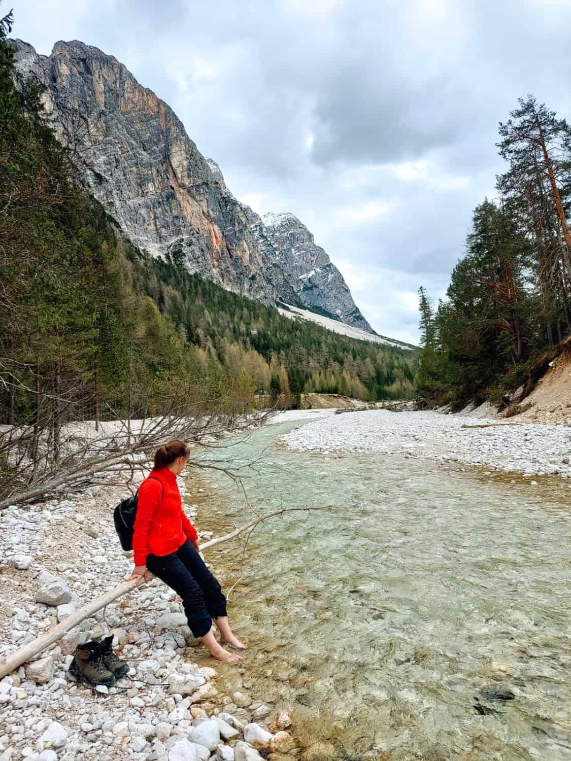 Dolomites In April: Perfect 2-Day Itinerary + What To Expect 55 A person in a red jacket and black pants sits on a log by a clear, rocky stream, dipping their feet in the water. Pine trees and a snowy mountain are in the background under a cloudy sky.