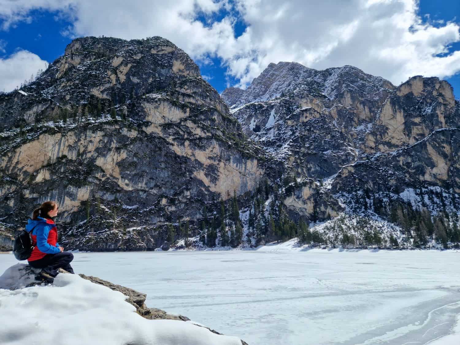 Dolomites In April: Perfect 2-Day Itinerary + What To Expect 54 A person in a red and blue jacket sits on a rock, overlooking a frozen, snow-covered lake. Majestic, snow-dusted mountains rise in the background under a partly cloudy sky.