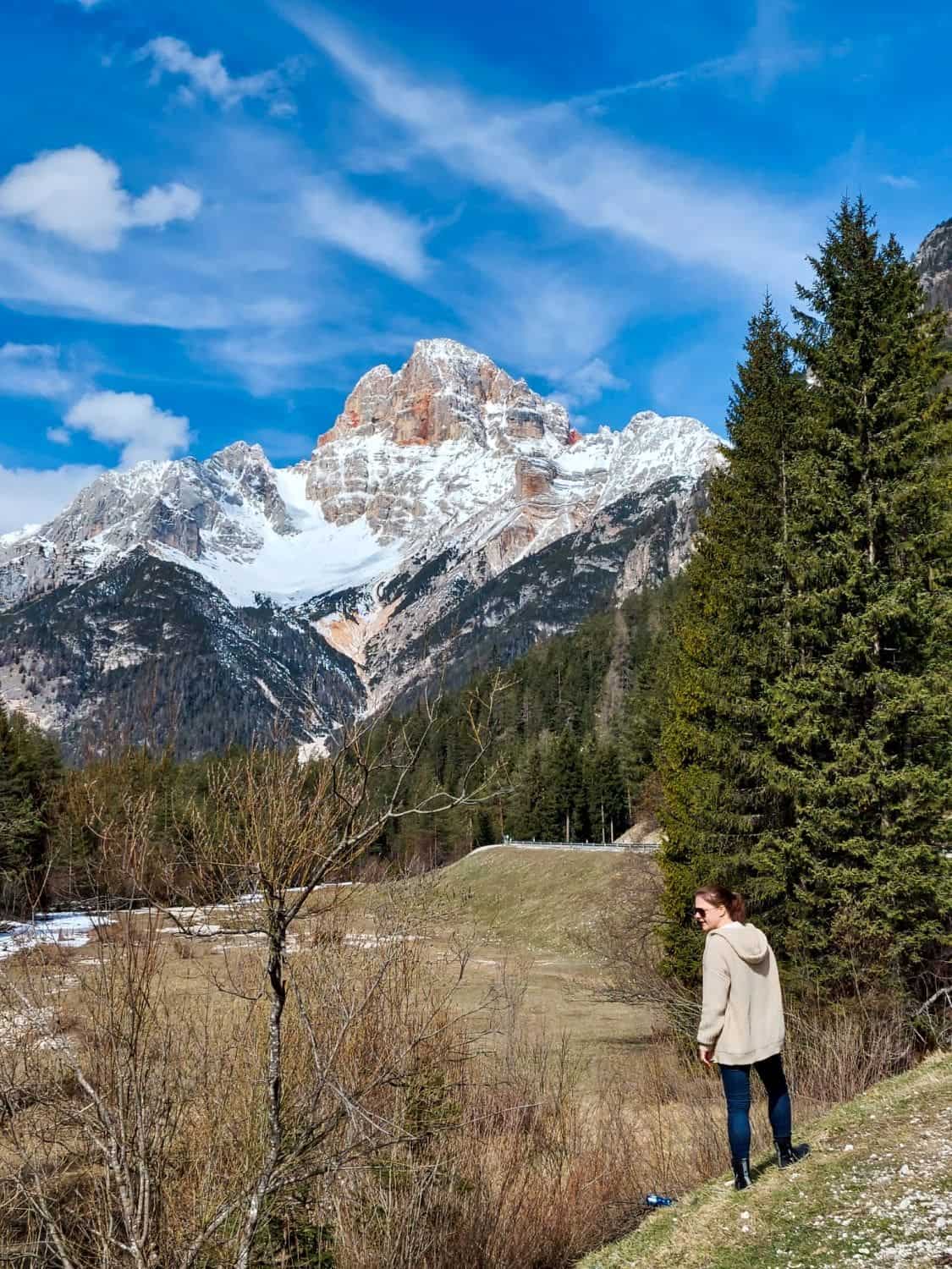 Dolomites In April: Perfect 2-Day Itinerary + What To Expect 48 A person stands on a grassy hill, wearing a light jacket, gazing at a mountainous landscape. Snow-capped peaks rise under a partly cloudy blue sky, surrounded by pine trees. Some snow patches are visible on the ground.