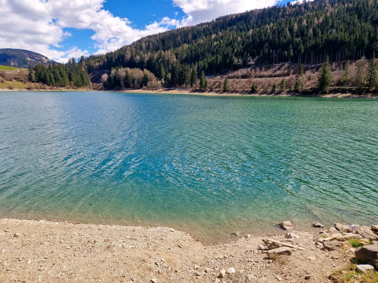 Dolomites In April: Perfect 2-Day Itinerary + What To Expect 13 A serene lake with clear turquoise water bordered by a rocky shore. Evergreen trees line the surrounding hills, under a partly cloudy blue sky. The landscape reflects a peaceful natural setting.