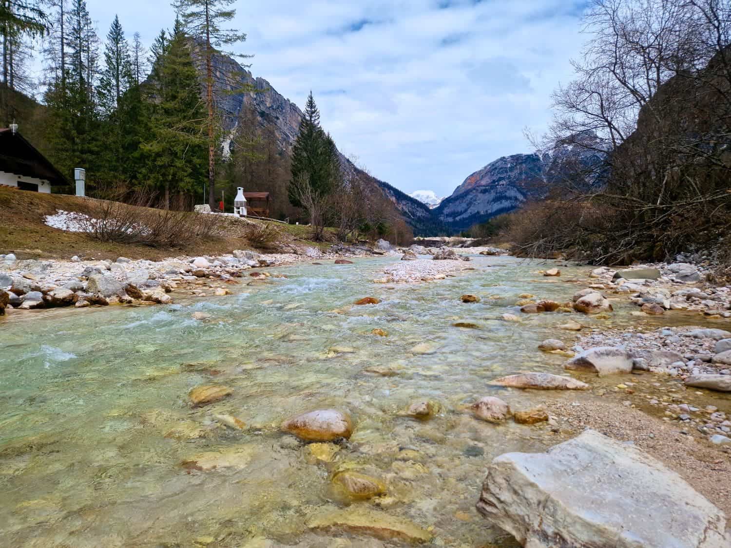 Dolomites In April: Perfect 2-Day Itinerary + What To Expect 29 A clear, shallow stream flows through a rocky riverbed, surrounded by leafless trees and evergreen forests. Snow-capped mountains rise in the background under a cloudy sky, creating a serene, natural landscape.