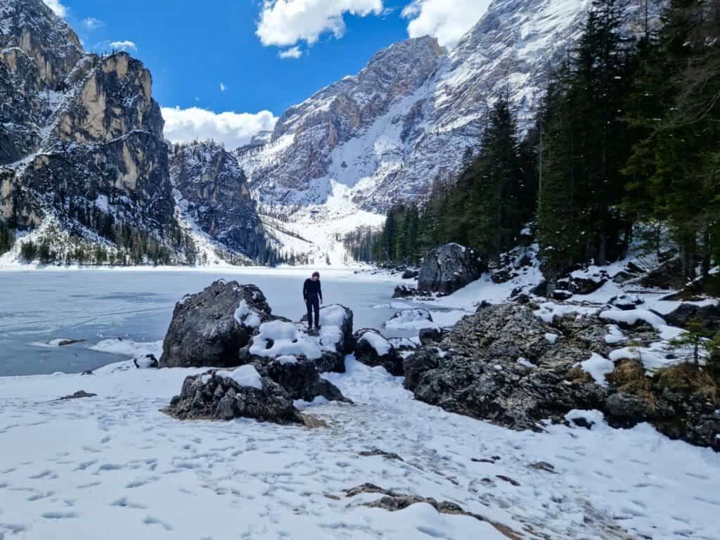 Dolomites In April: Perfect 2-Day Itinerary + What To Expect 16 A person stands on snow-covered rocks by a partially frozen lake surrounded by snow-dusted mountains and pine trees under a cloudy blue sky.
