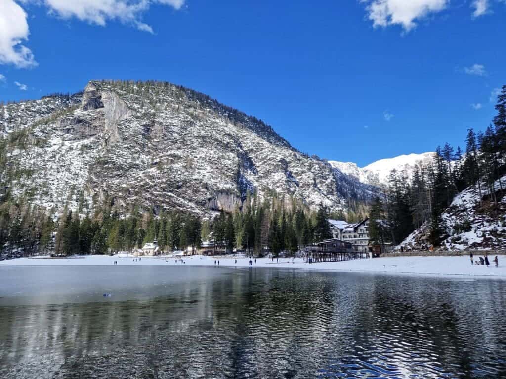 Dolomites In April: Perfect 2-Day Itinerary + What To Expect 14 Snow-covered lake with surrounding mountains and pine trees under a clear blue sky. Small groups of people are walking on the frozen surface. A lodge is visible near the edge of the lake.
