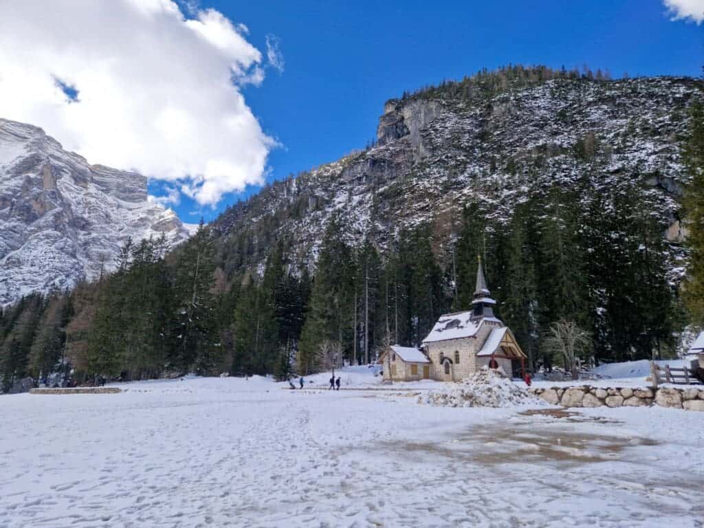 Dolomites In April: Perfect 2-Day Itinerary + What To Expect 15 A small chapel with a pointed roof sits amidst a snowy landscape. Tall evergreen trees surround the area, and a rocky mountain rises in the background. People are walking near the chapel, under a partly cloudy blue sky.