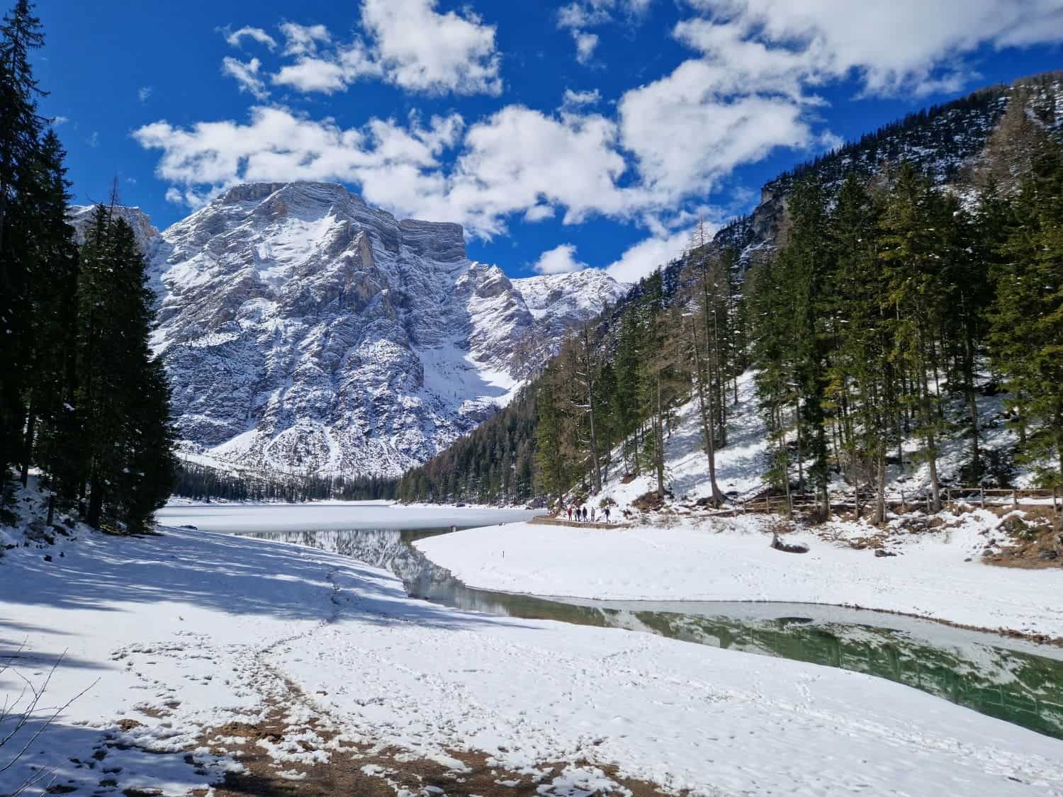 Dolomites In April: Perfect 2-Day Itinerary + What To Expect 8 Snow-covered landscape with a partially frozen lake surrounded by evergreen trees. A rugged mountain range rises in the background under a partly cloudy blue sky. A path leads towards the water's edge.