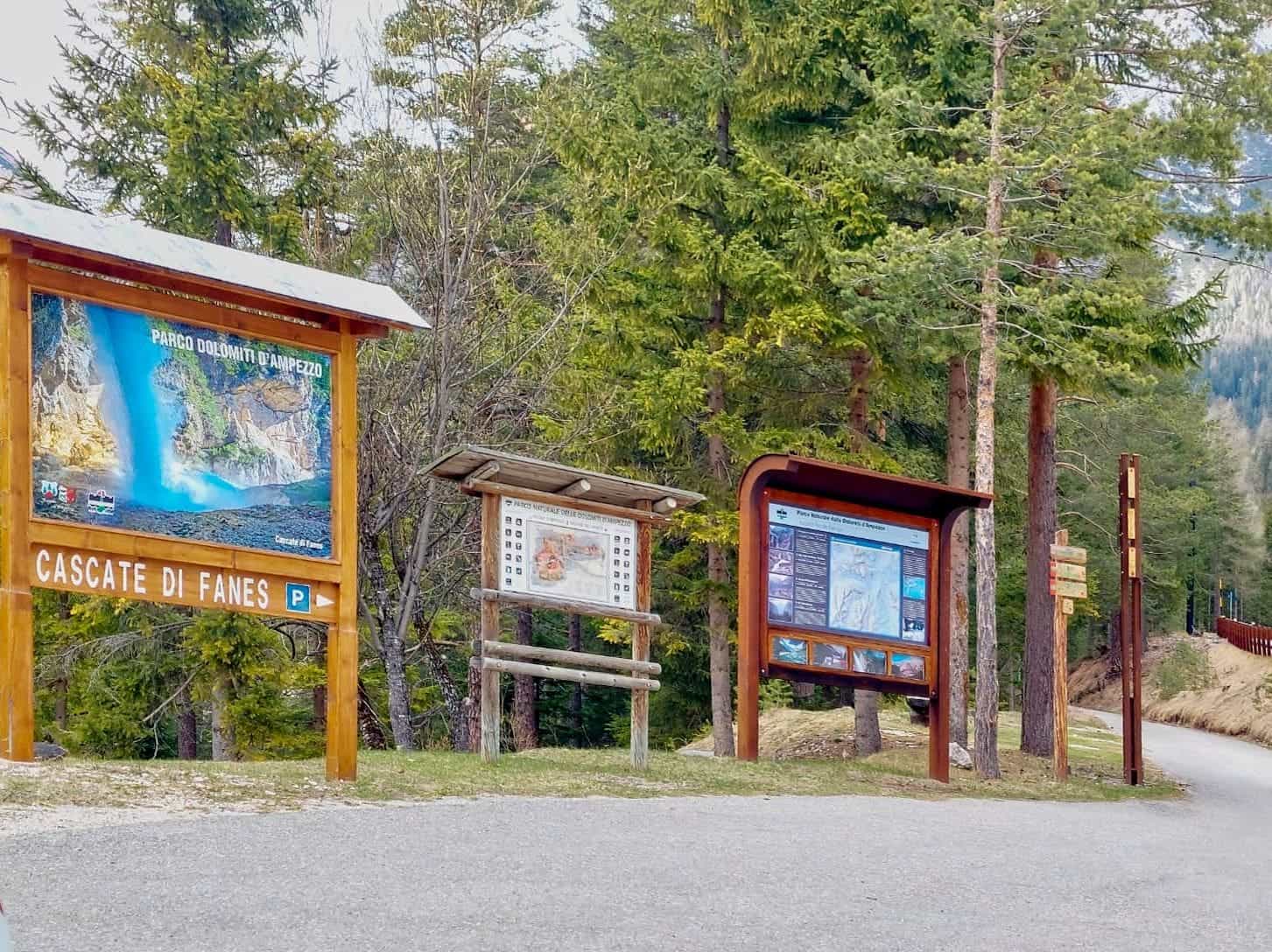 Dolomites In April: Perfect 2-Day Itinerary + What To Expect 37 Signs and information boards at the entrance of a wooded area, including a large map labeled "Cascate di Fanes" on the left. Tall pine trees and a walking path are visible in the background.