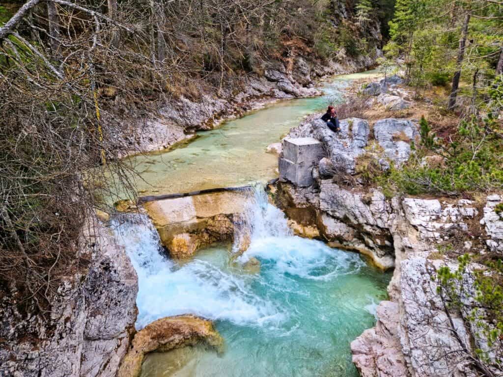 Cool Things To Do In The Dolomites: From Hiking to Flying 17 A person sits on a rock ledge near a cascading turquoise stream surrounded by rugged rock formations and dense forest. The water flows energetically, creating small rapids as it winds through the serene natural landscape.