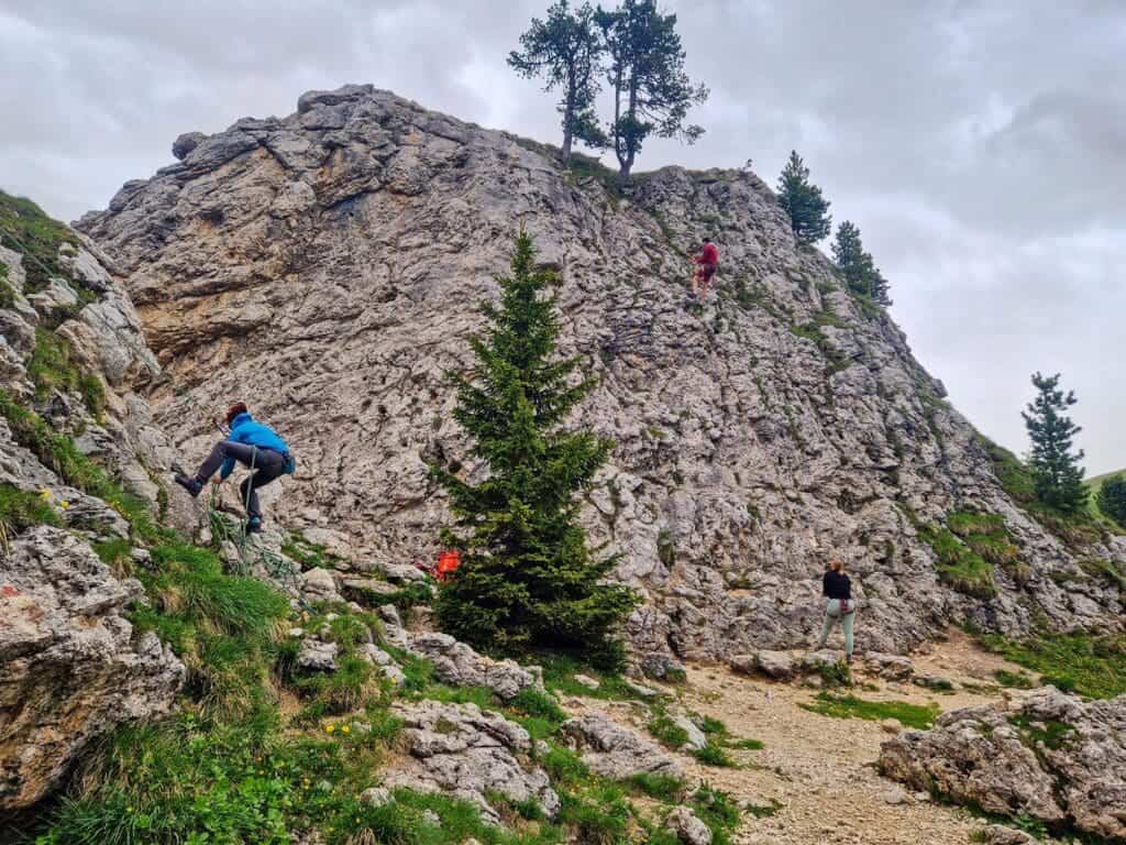 Three people rock climbing on a rugged, rocky hillside with some greenery. A few trees are scattered along the slope. The sky is cloudy, adding a dramatic backdrop to the scene.