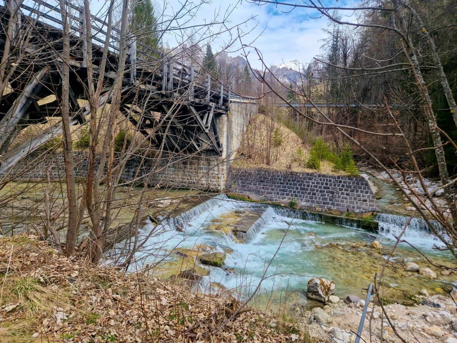 Dolomites In April: Perfect 2-Day Itinerary + What To Expect 32 A rustic wooden bridge stretches over a rocky stream with small waterfalls. Bare trees surround the scene, and distant mountains rise against a cloudy sky. Fallen leaves cover the ground, suggesting late autumn or early spring.