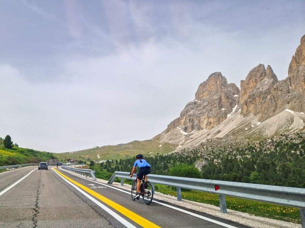 A cyclist wearing a blue shirt rides along a road surrounded by mountainous terrain. The road features distinct bike and car lanes. In the background, large rocky peaks rise under a partly cloudy sky.