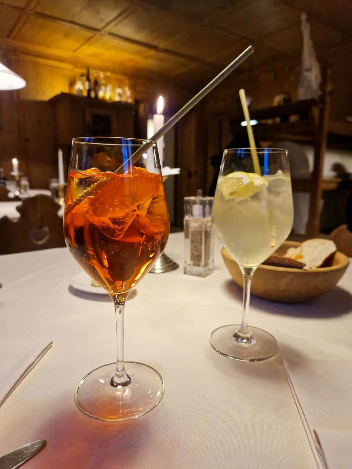 Dolomites In April: Perfect 2-Day Itinerary + What To Expect 4 Two glasses of cocktails on a table with a white tablecloth. One glass contains an orange drink with ice and an orange slice; the other a light-colored drink with ice and a lemon slice. A bowl of bread is in the background, with a cozy, dimly-lit setting.