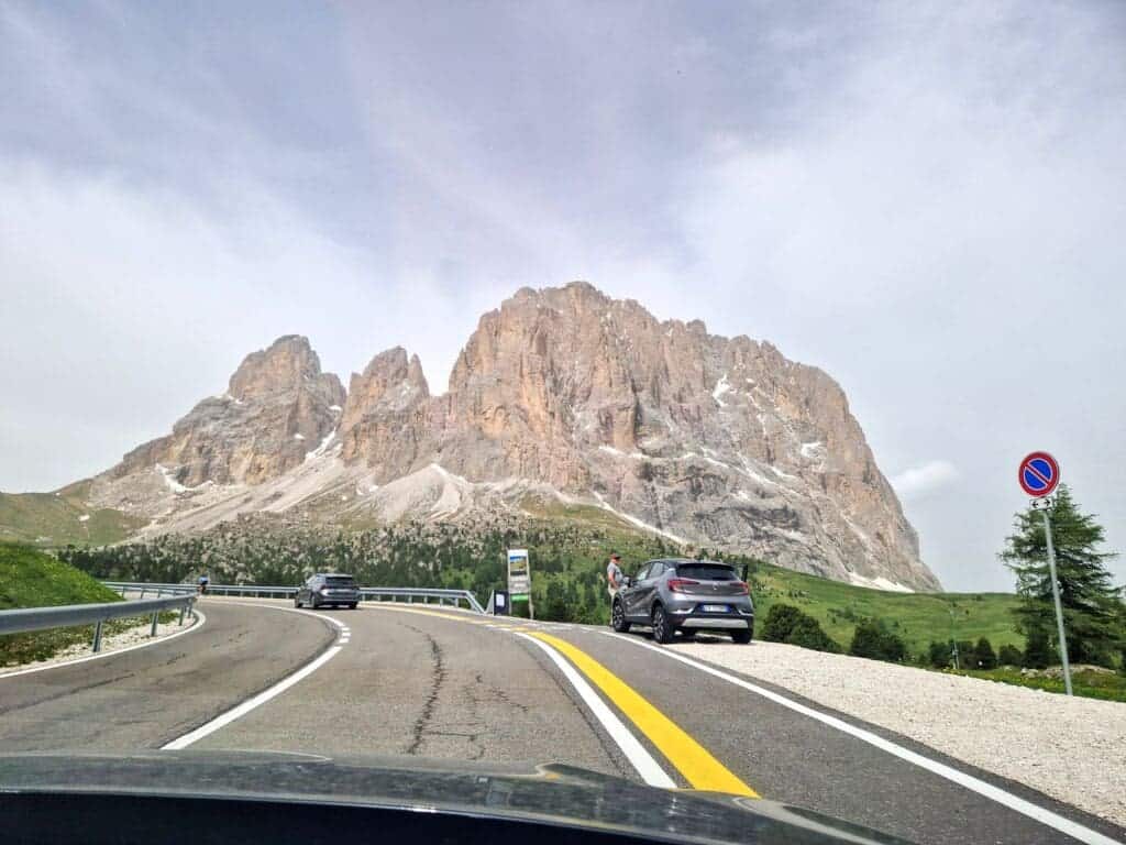 A winding road leads to majestic, rocky mountains under a cloudy sky. Cars are parked along the roadside, with one person visible near a parked car. Lush green grass contrasts the rugged mountain terrain. A no-parking sign is also visible.