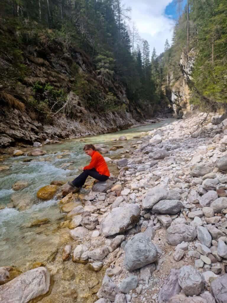 Dolomites In April: Perfect 2-Day Itinerary + What To Expect 44 A person in a red jacket sits on a rock by a rocky riverbank surrounded by forested hills. The water is clear, and the sky is partly cloudy. The scene conveys a peaceful, natural setting.