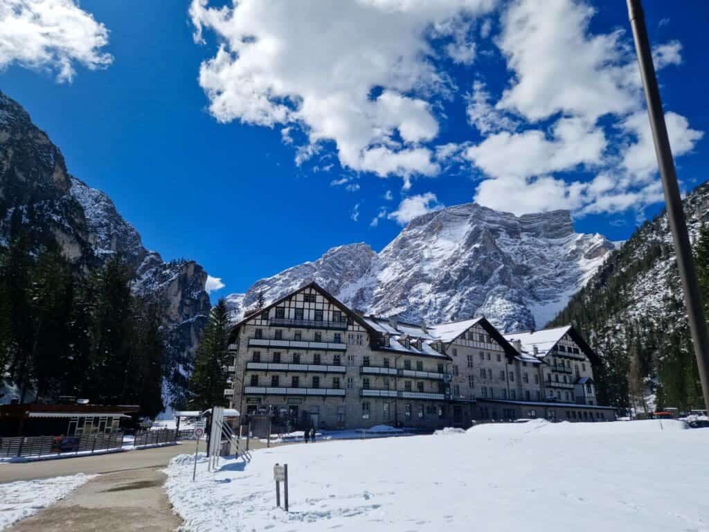 Dolomites In April: Perfect 2-Day Itinerary + What To Expect 5 A large, alpine-style hotel stands amidst a snowy landscape with towering mountains in the background. The sky is bright blue with scattered fluffy clouds. Pine trees flank the building, adding to the picturesque, serene setting.