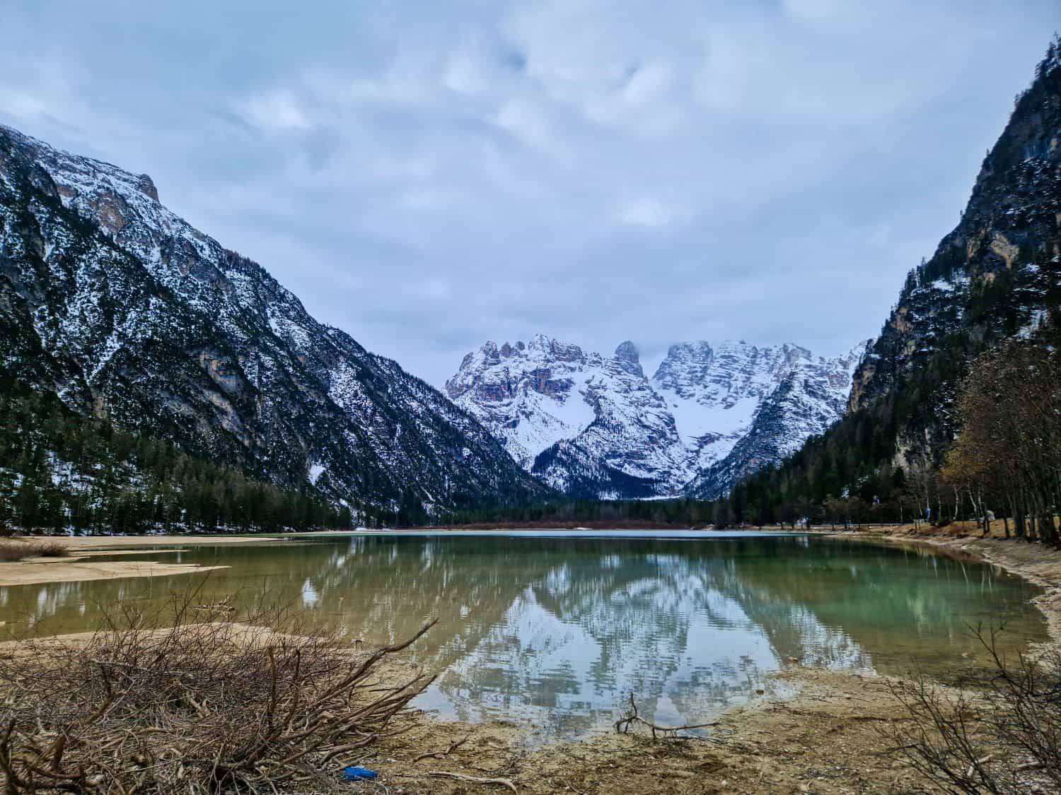 Dolomites In April: Perfect 2-Day Itinerary + What To Expect 2 A serene mountain lake with clear reflections of snow-capped peaks and surrounding forests under a cloudy sky. The landscape is framed by rugged mountains on both sides, with a mix of bare and evergreen trees lining the shore.